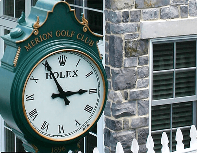 We see a large, green, Rolex-branded clock displaying the time as 2:55 PM in front of a grey manufactured stone veneer corner of the Classroom and Driving range at the US Open Golf Tournament held at the Merion Country Club.